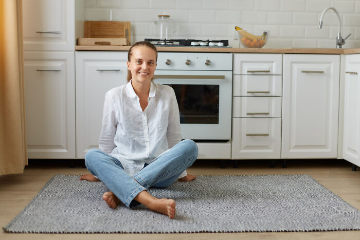 Young woman sitting cross-legged on a rug in a modern kitchen reflecting on childhood habits and normal behavior.