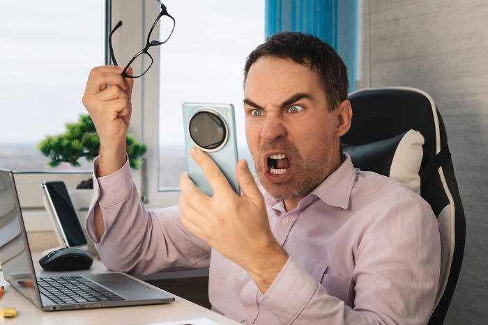 Angry man holding glasses and phone, reacting with frustration while sitting at a desk showing signs of cheating suspicion.