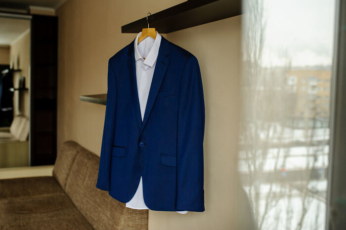 Blue suit jacket and white shirt hanging on a wooden hanger indoors near a window, illustrating formal Thanksgiving gathering attire.