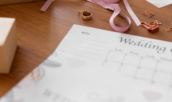 Wedding planner with rings and pink ribbons on wooden table symbolizing bride wants to upgrade honeymoon at bridesmaid’s expense.