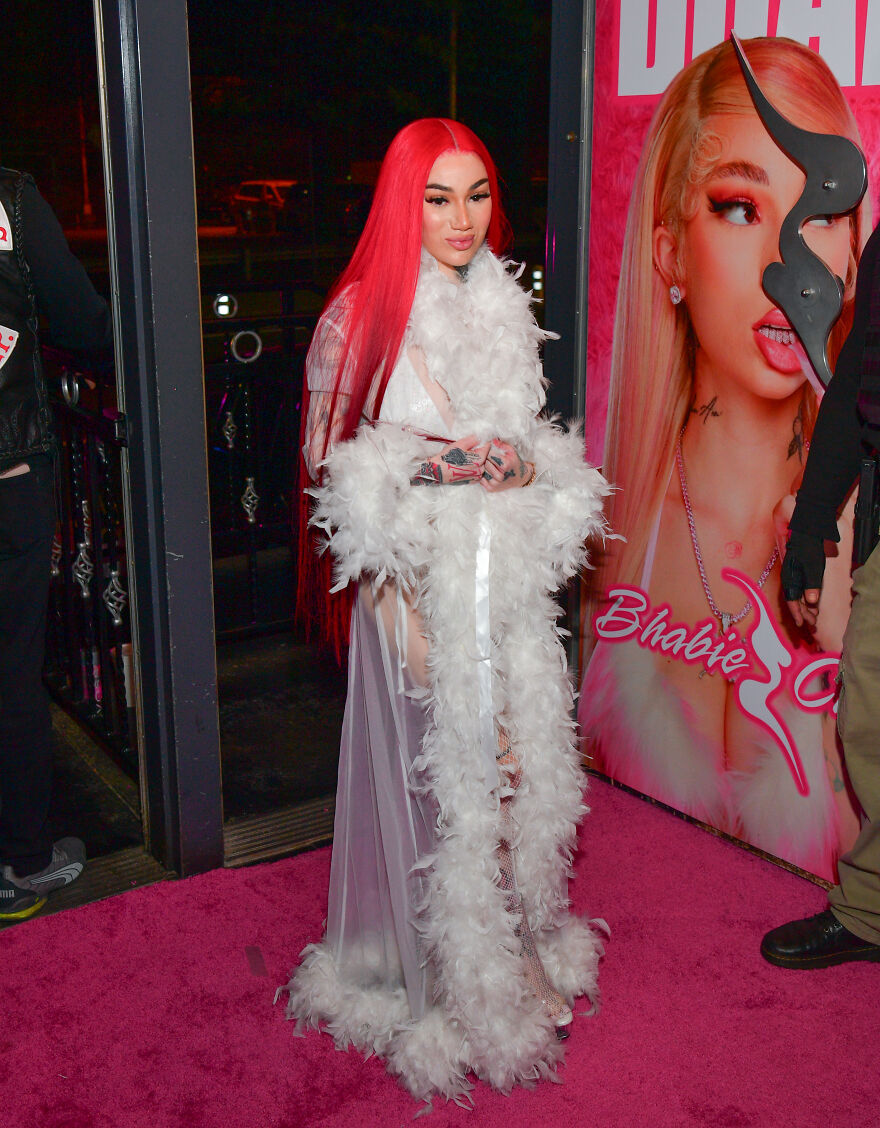 Bhad Bhabie with long red hair in a white feathered outfit posing on a pink carpet for members-only fan pages event.