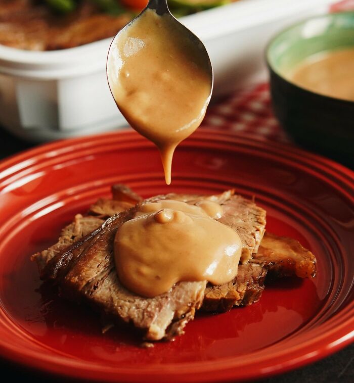 Spoon drizzling thick gravy over sliced roast beef on a red plate, highlighting popular Thanksgiving mashed potatoes debate.