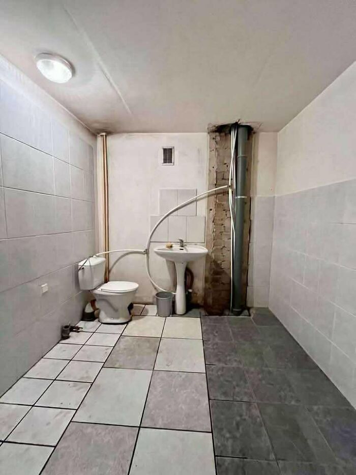 Small bathroom in a real estate listing with mismatched tiles, exposed pipes, and an unfinished wall area under a ceiling light.