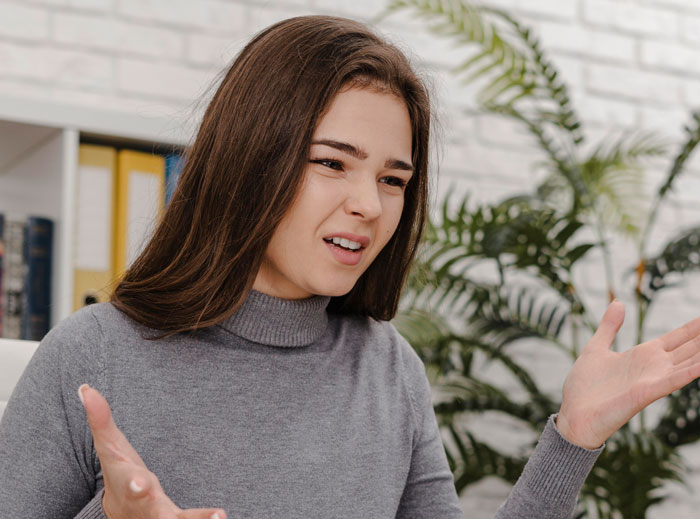 Young woman in gray sweater expressing frustration indoors, illustrating bride wanting to upgrade honeymoon at bridesmaid’s expense concept.