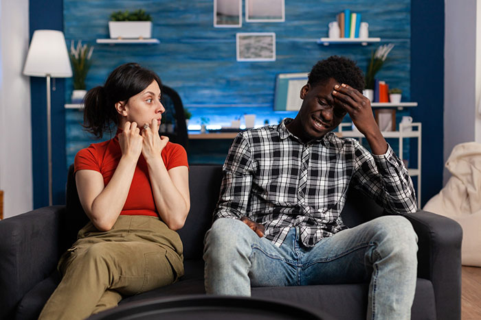 Man looking hurt and frustrated sitting on couch while wife avoids eye contact, showing family and work priorities conflict. Man looking hurt and frustrated sitting on couch while wife avoids eye contact, showing family and work priorities conflict.