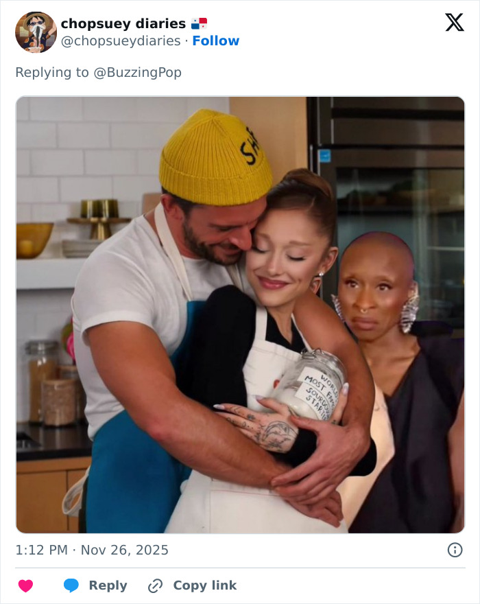 Ariana Grande and Jonathan Bailey sharing an intimate moment in a kitchen while another person looks on. Ariana Grande and Jonathan Bailey sharing an intimate moment in a kitchen while another person looks on.
