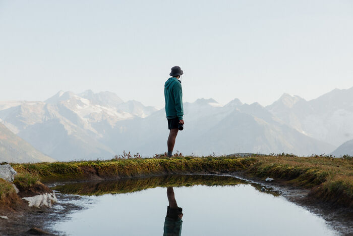 Person standing near a mountain lake reflecting snowy peaks, symbolizing a journey to find purpose and direction. Person standing near a mountain lake reflecting snowy peaks, symbolizing a journey to find purpose and direction.