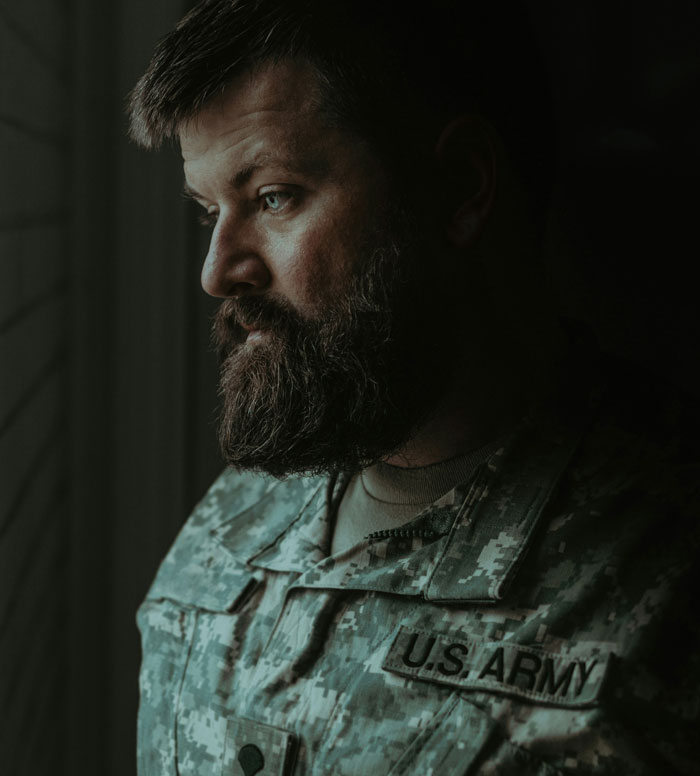 A pensive man in U.S. Army uniform looking out a window, illustrating frustration related to HOA rules and regulations.