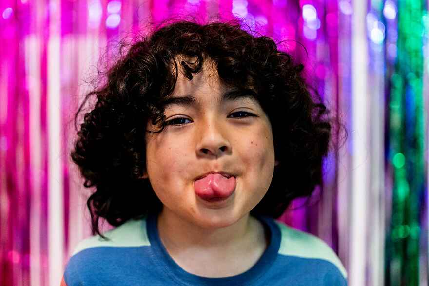 Child with curly hair sticking out tongue playfully in front of colorful shiny backdrop illustrating tongue twisters challenge.