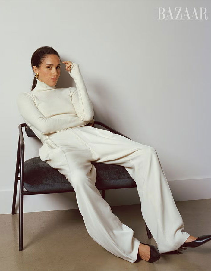 Woman in white turtleneck and trousers seated on chair, showcasing Meghan Markle Harper’s Bazaar moment style and elegance.