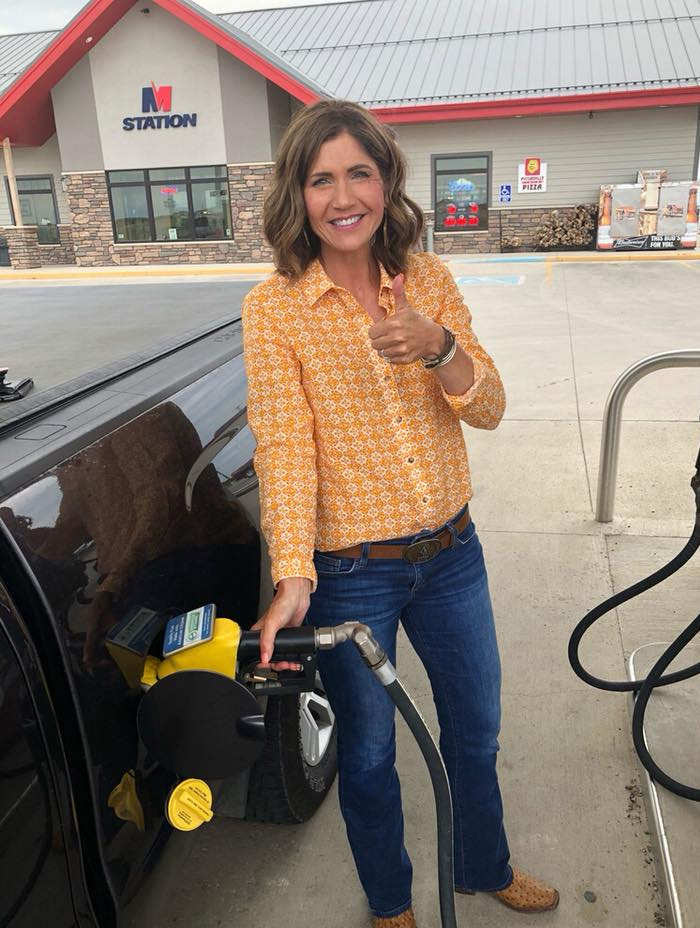 Kristi Noem smiling while pumping gas at a station, wearing a yellow patterned shirt and blue jeans outdoors.