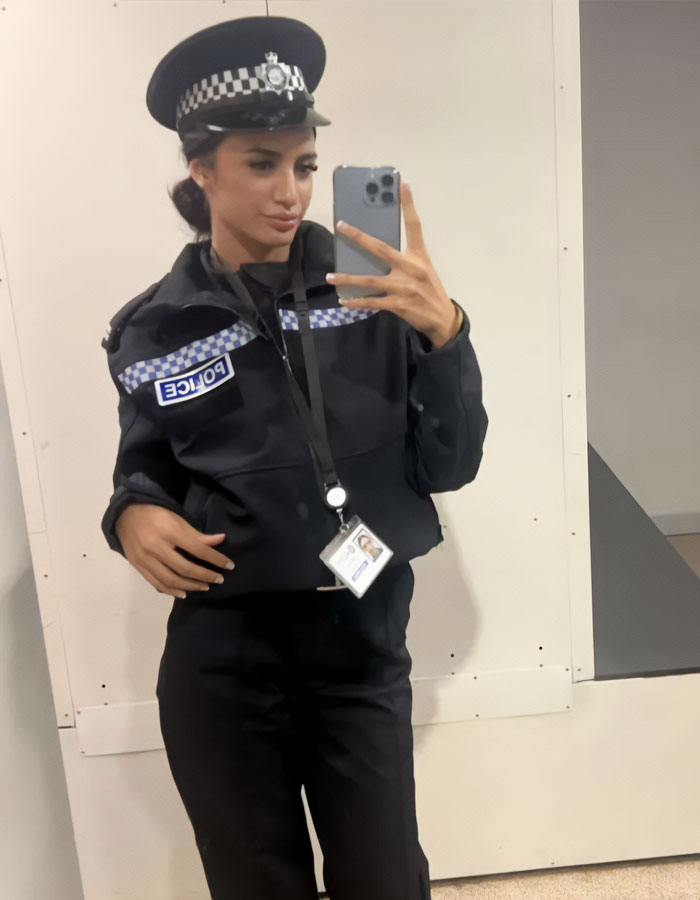 Female trainee police officer in uniform taking a mirror selfie with badge and cap in an indoor setting.