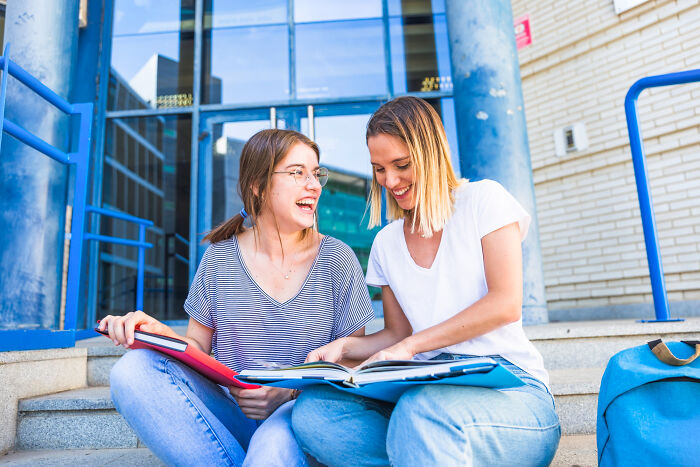 Two young women sitting on outdoor steps, smiling and sharing books, symbolizing people who disappeared to start new lives.