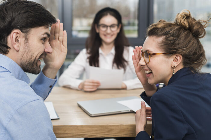 Two workers sharing unprofessional things done, laughing and whispering during a casual office meeting with a third person.