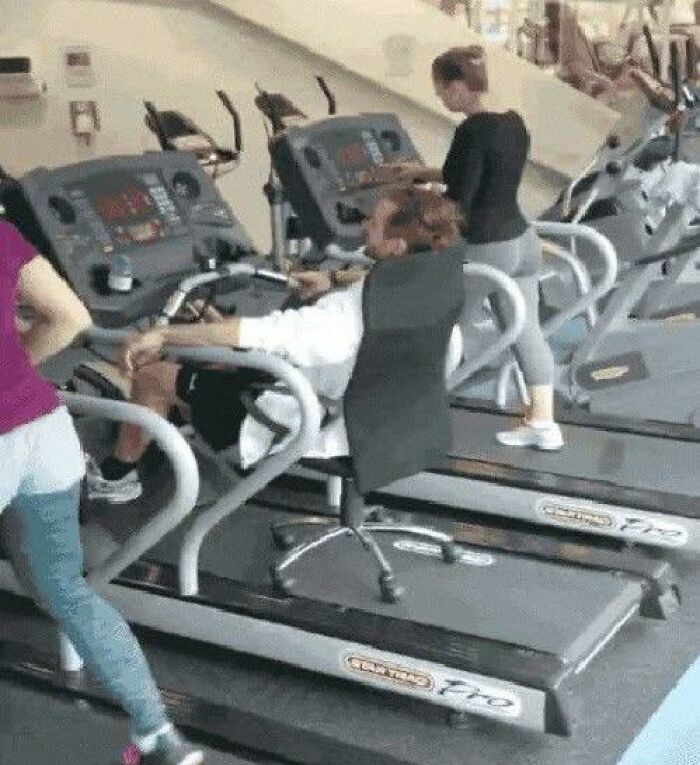 Man sitting on an office chair going hard on a treadmill in a gym while others exercise on regular treadmills nearby.