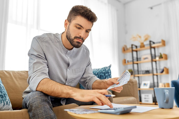 Man using calculator and holding bills on couch, reflecting on plans with uneasy sleeping arrangements involved.