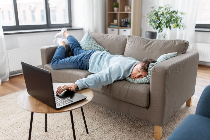 Man lying face down on sofa exhausted, reaching for laptop, depicting infuriating work routine experiences.