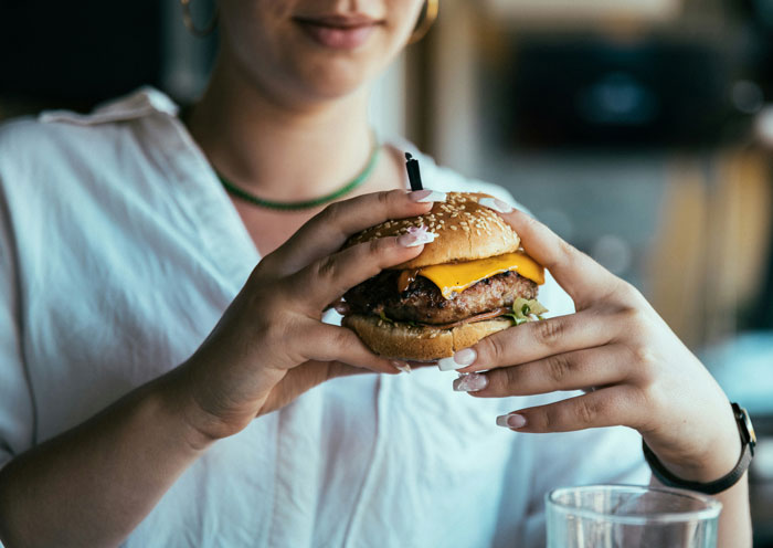 Woman eating a cheeseburger she shouldn’t have, coworker paying the price in a casual dining setting.