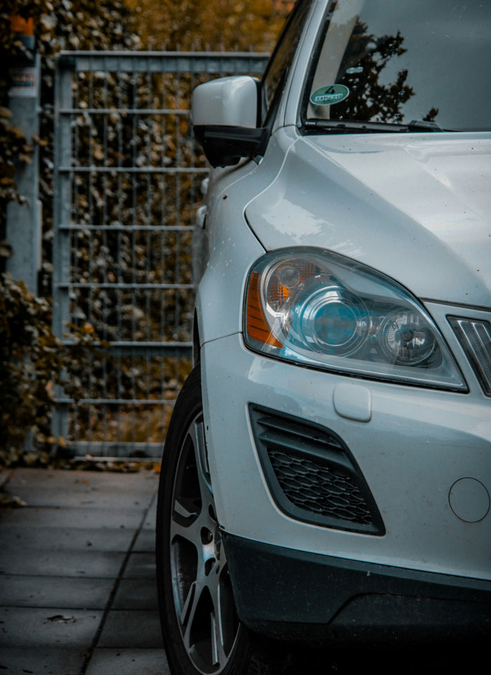 Close-up of a white car with headlight on, parked near a metal gate and autumn foliage, illustrating frustration with people.