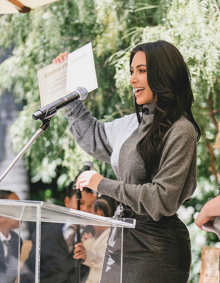 Kim Kardashian speaking at podium, holding certificate, related to her excuses for failing the bar exam so far.