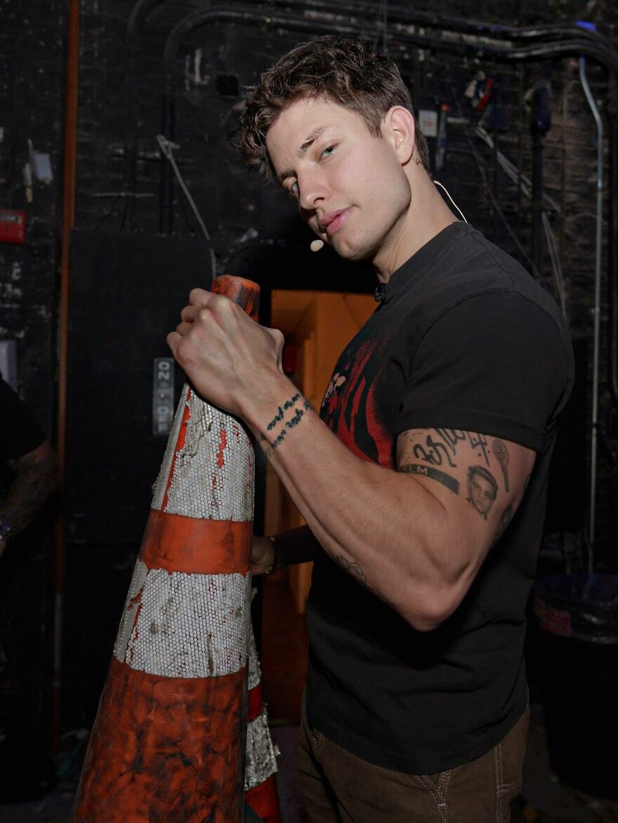 Matt Rife holding an orange and white traffic cone backstage, showcasing tattoos and wearing a black shirt. Matt Rife holding an orange and white traffic cone backstage, showcasing tattoos and wearing a black shirt.