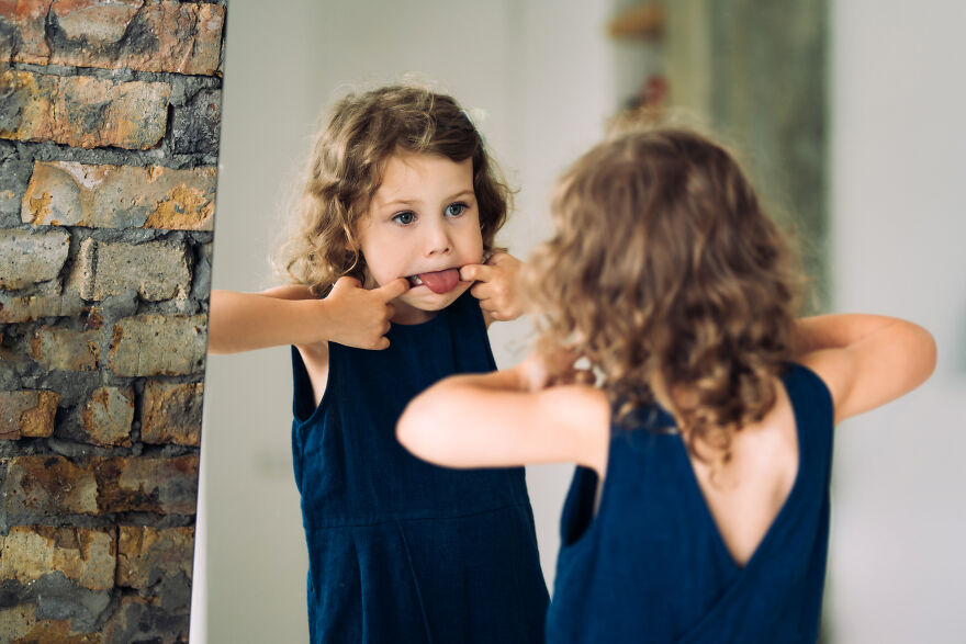 Young girl in a blue dress making a funny face in front of a mirror, embodying tongue twisters challenge playfully.