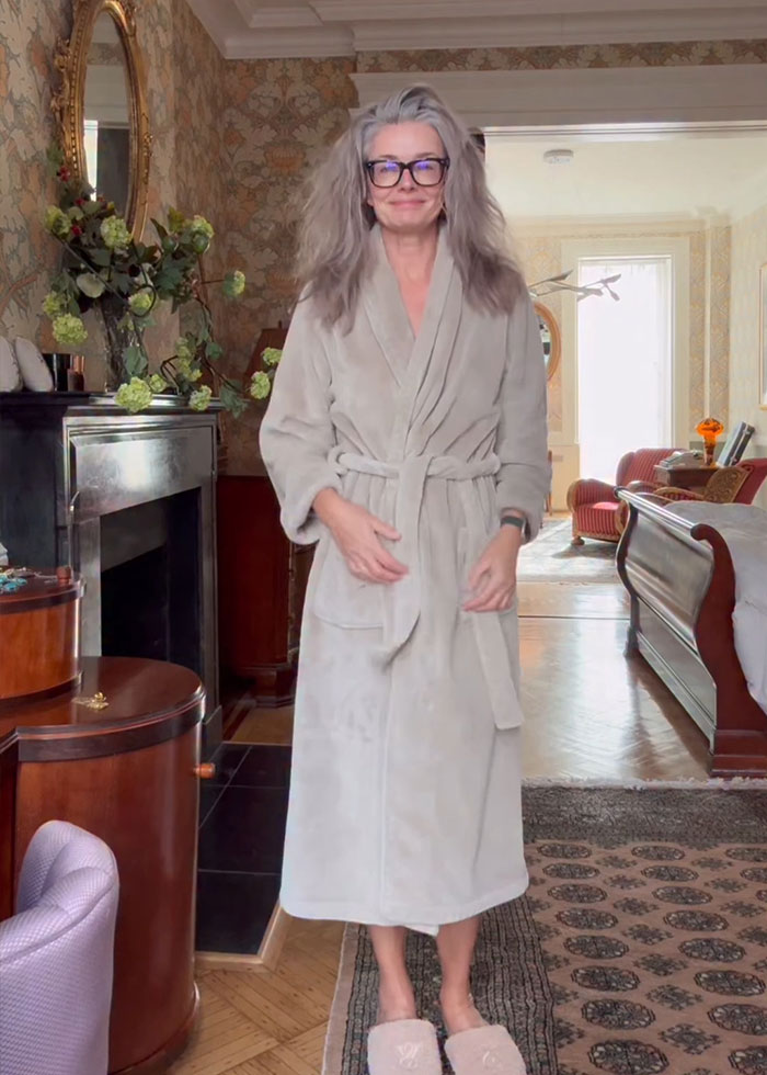 Paulina Porizkova, 60, in a beige robe and slippers, embracing her perfect imperfections in a cozy home setting.