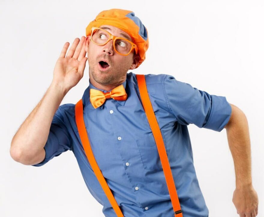 A Blippi dressed as Blippi wearing bow tie, orange suspenders, and glasses, posing with hand near ear on white background A Blippi dressed as Blippi wearing bow tie, orange suspenders, and glasses, posing with hand near ear on white background