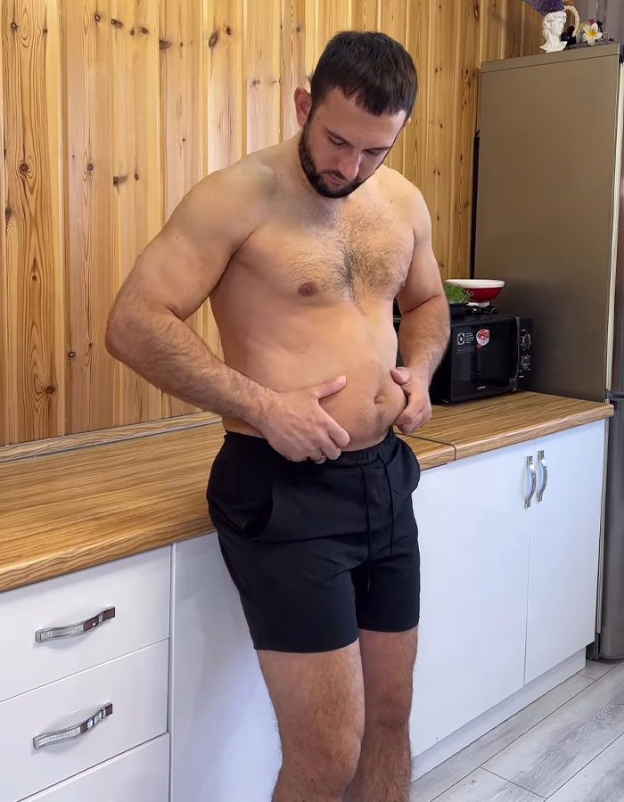 Fitness coach shirtless in kitchen, examining his stomach while wearing black shorts, wooden panel background. Fitness coach shirtless in kitchen, examining his stomach while wearing black shorts, wooden panel background.