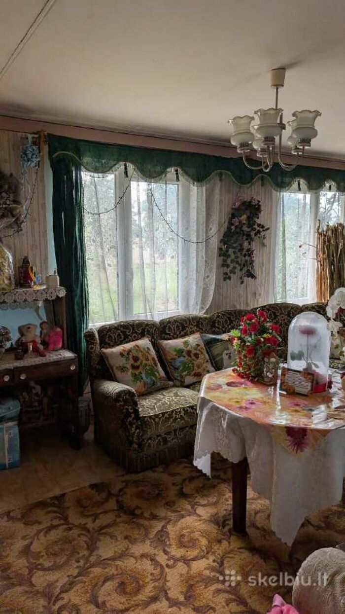 Vintage living room with floral cushions, patterned carpet, and lace curtains in a wild real estate listing interior.