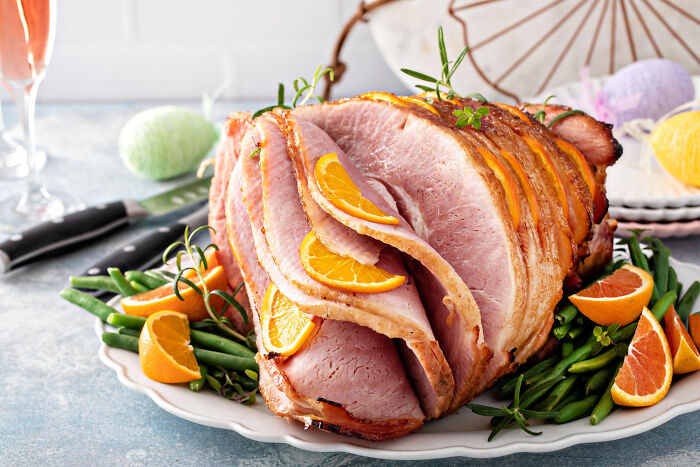Spiral sliced ham garnished with orange slices and herbs served with green beans on a white platter.