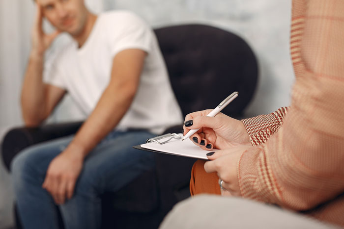 Man sitting on couch looking troubled during therapy session with counselor writing on clipboard about autistic brother behavior.