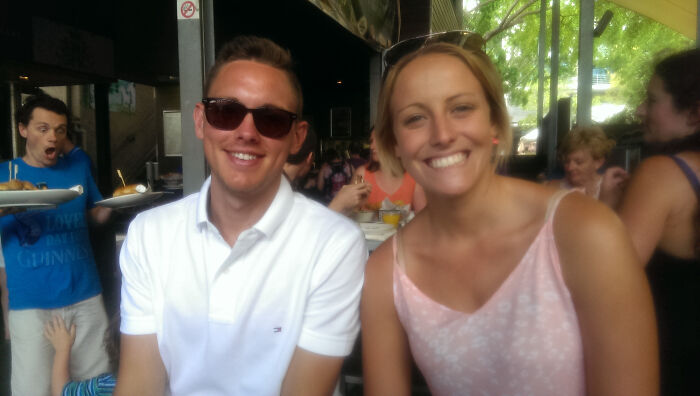 Two people smiling at a cafe with a man in the background photobombing and making a surprised face.