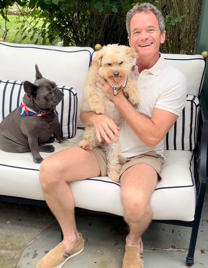 Neil Patrick Harris sitting on a white bench outdoors holding a dog with another dog beside him, smiling.