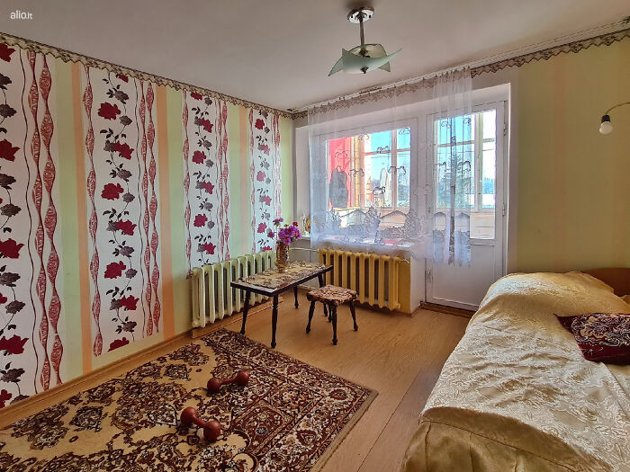 Cozy vintage bedroom with floral wallpaper, ornate rug, small table, and large window in wild real estate listing photo.