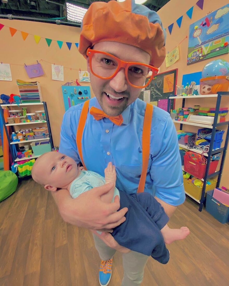 A Blippi wearing orange bow tie and glasses holding a baby in a colorful, playful room with educational decorations. A Blippi wearing orange bow tie and glasses holding a baby in a colorful, playful room with educational decorations.