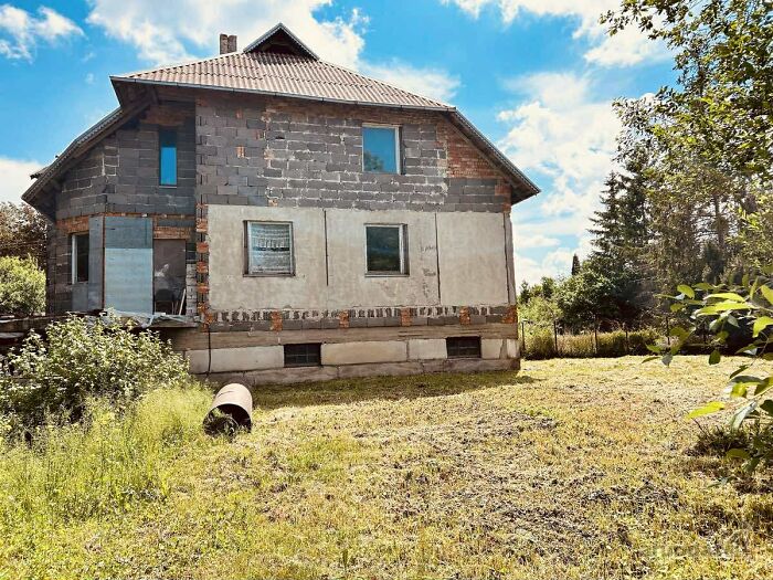 Unfinished real estate listing featuring a partially constructed house with exposed brick and overgrown yard under blue sky.