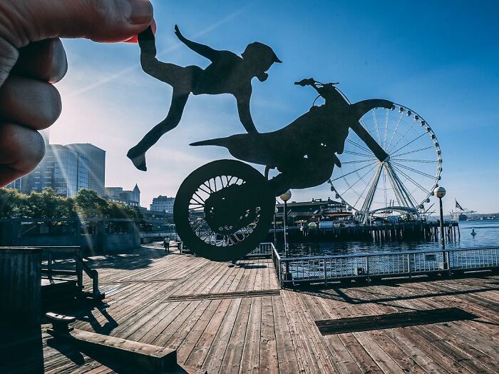 Hand holding paper cutouts of a motorcyclist and a person, creatively interacting with a waterfront Ferris wheel scene.