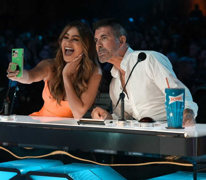 Simon Cowell leaning forward at judging panel with woman in orange dress taking selfie during show. Simon Cowell leaning forward at judging panel with woman in orange dress taking selfie during show.