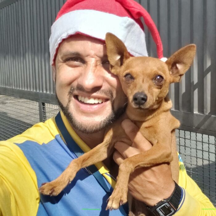 Mailman wearing a red hat smiling and holding a small dog while standing outdoors near a fence on his route.