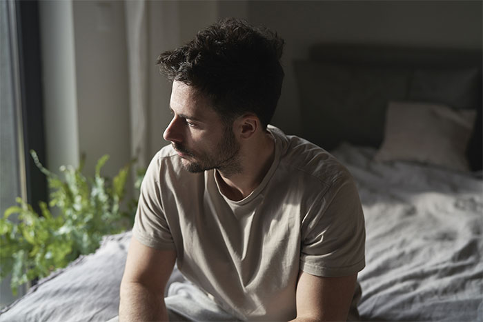 Man sitting on bed looking away thoughtfully, reflecting on girlfriend kissing another woman in front of friends. Man sitting on bed looking away thoughtfully, reflecting on girlfriend kissing another woman in front of friends.
