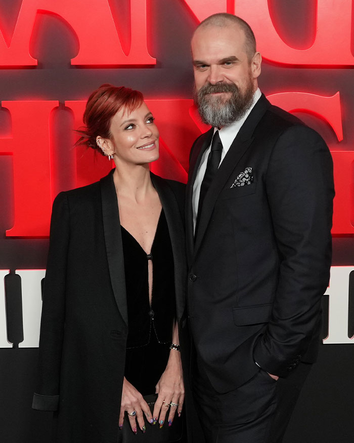David Harbour and Stranger Things co-star smiling at event with red Stranger Things backdrop behind them. David Harbour and Stranger Things co-star smiling at event with red Stranger Things backdrop behind them.