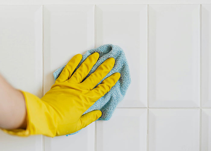 Hand wearing yellow glove wiping white tile wall with blue cloth showing genius cleaning hacks inspired by laziness.