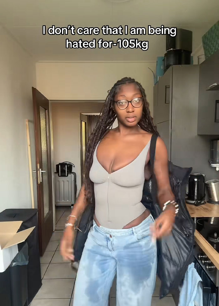 Woman demonstrating trick to hide loose skin on her belly after massive weight loss while standing in a kitchen. Woman demonstrating trick to hide loose skin on her belly after massive weight loss while standing in a kitchen.