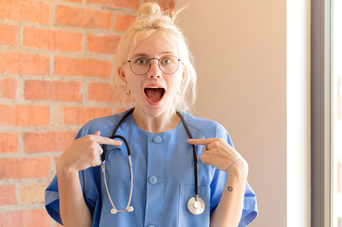 Young female healthcare worker in blue scrubs pointing at her stethoscope with a funny surprised expression.
