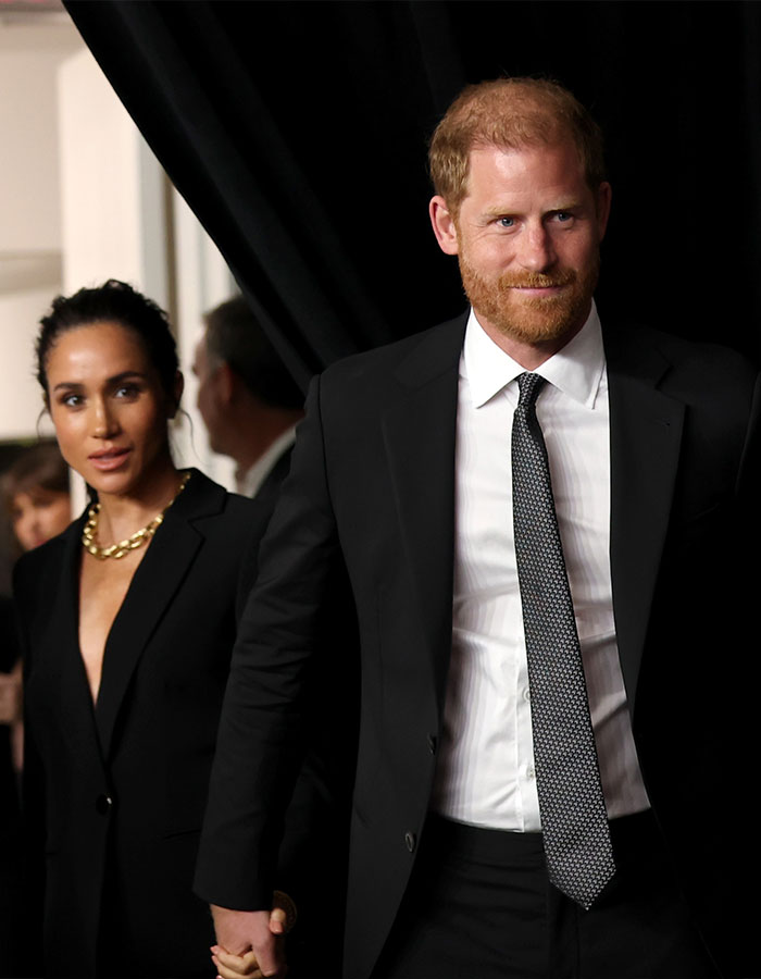 Meghan Markle and Prince Harry dressed in black, attending an event, with Meghan wearing a gold chain necklace.