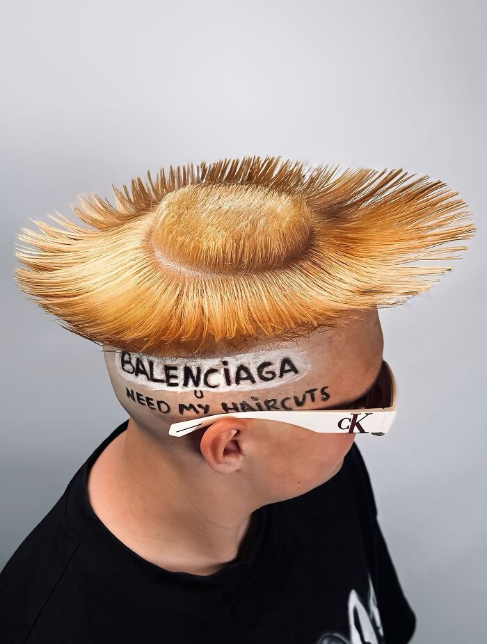 Young man with bold barber haircut styled in upright spikes wearing white sunglasses and a black shirt on a gray background