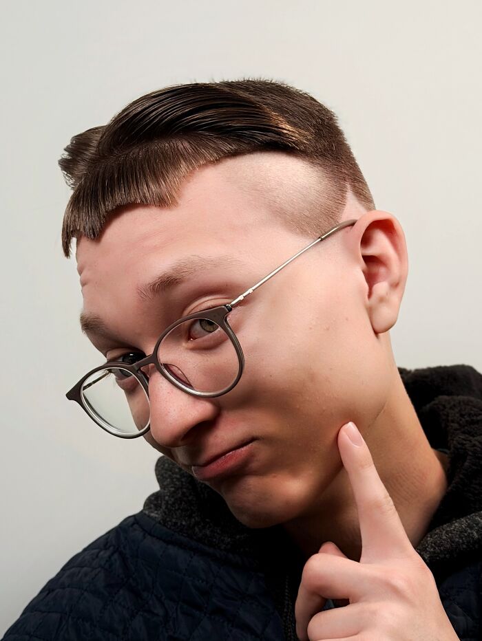 Young man with bold barber haircut featuring sharp lines and artistic styling wearing glasses and a dark hoodie against a white background.
