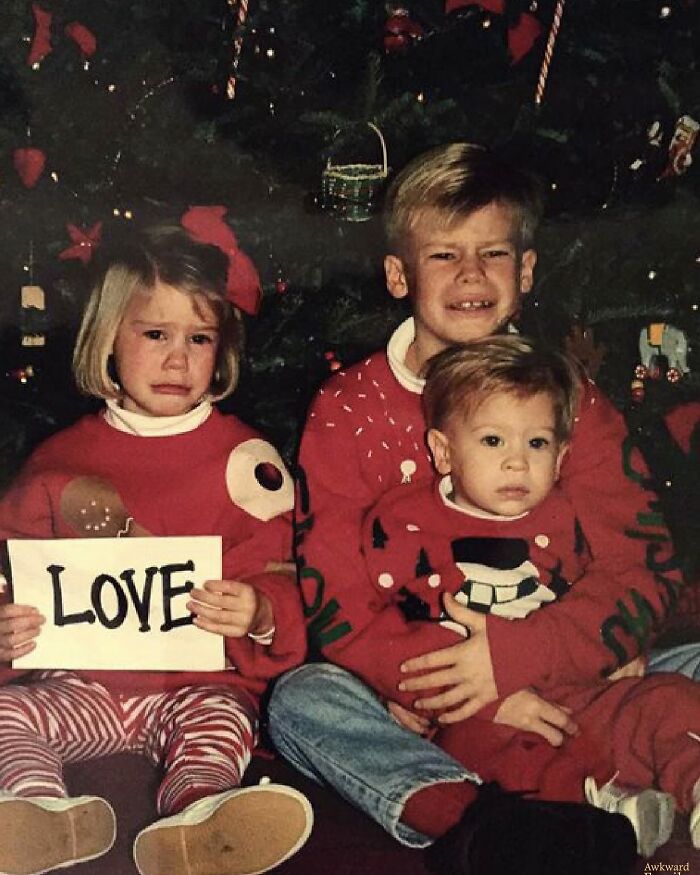 Three kids in awkward studio pics wearing festive sweaters sitting in front of a Christmas tree, one holding a love sign.