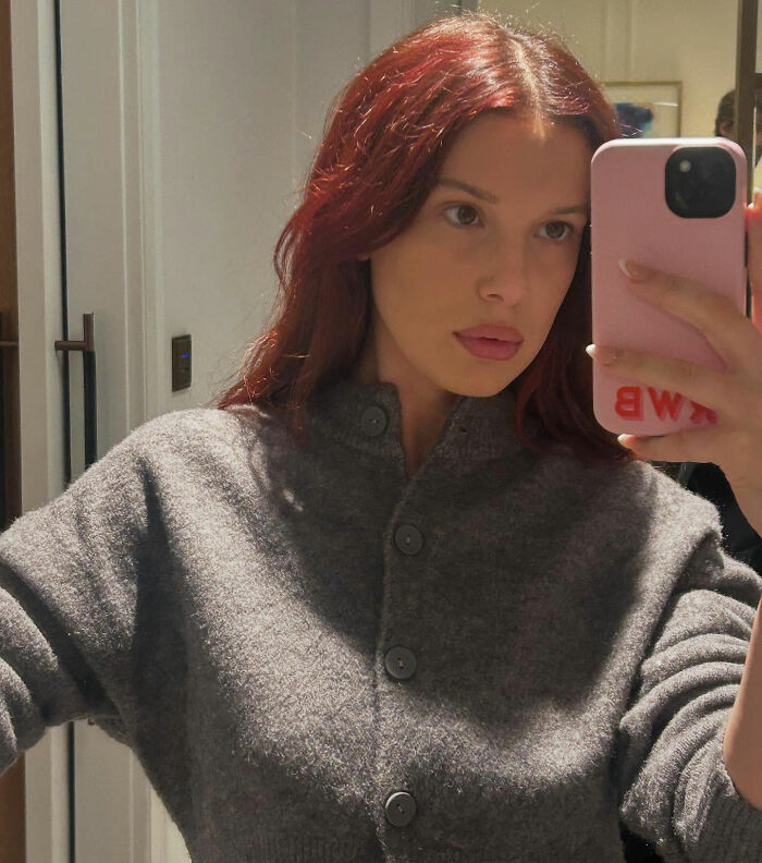 Millie Bobby Brown taking a mirror selfie with red hair and a grey buttoned sweater holding a pink phone case. Millie Bobby Brown taking a mirror selfie with red hair and a grey buttoned sweater holding a pink phone case.
